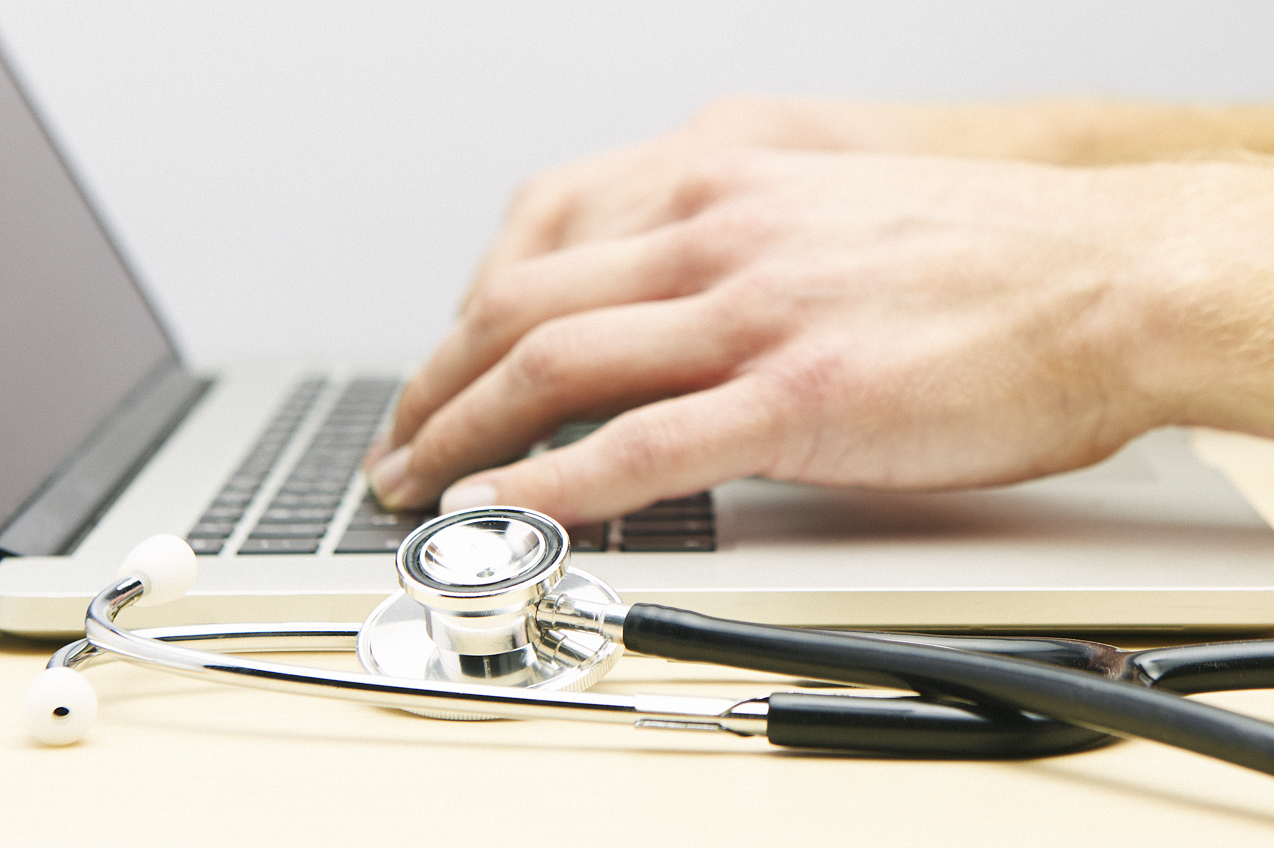Hands typing on a laptop with a stethoscope in the foreground.